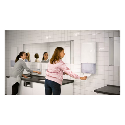 Deux femmes se tiennent dans une salle de bain avec des murs carrelés blancs et de grands miroirs. L'une utilise un lavabo sur le côté gauche, tandis que l'autre tire sans effort une serviette sans fin d'un distributeur Tork PeakServe® 552500 pour serviettes sans fin Elevation H5 de TORK – Essity Professional Hygiene Germany GmbH, situé sur le côté droit. Le plan de travail gris complète l'éclairage lumineux de cette salle de bain très fréquentée.