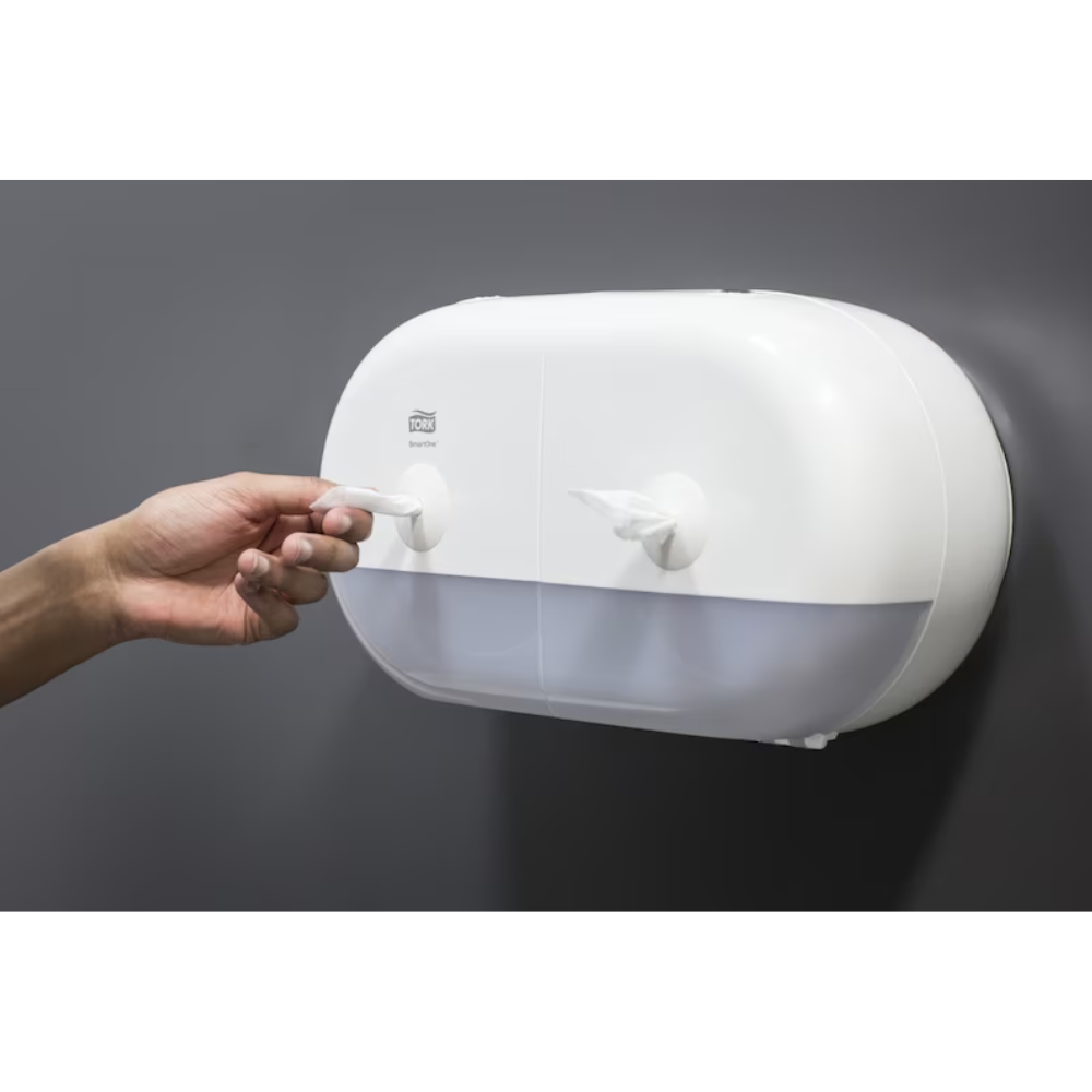Une personne tire une serviette en papier d'un élégant distributeur double rouleaux blanc Tork SmartOne® 682000 Mini pour papier toilette, qui dispose de deux compartiments et est fixé à un mur lisse et gris foncé.