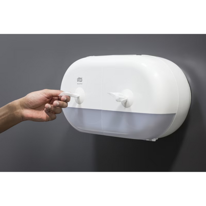 Une personne tire une serviette en papier d'un élégant distributeur double rouleaux blanc Tork SmartOne® 682000 Mini pour papier toilette, qui dispose de deux compartiments et est fixé à un mur lisse et gris foncé.