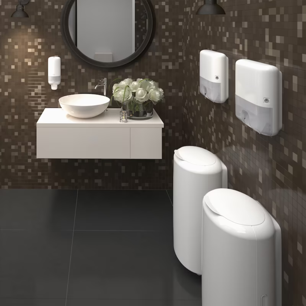 Une toilette publique moderne avec un mur de carreaux de mosaïque bruns dispose d'un miroir rond, d'un lavabo mural avec une cuvette blanche, de distributeurs de savon, du Tork Xpress® 552100 Mini Distributeur pour serviettes pliées H2 Elevation de TORK – Essity Professional Hygiene Germany GmbH et de deux poubelles cylindriques blanches. Un bouquet de fleurs blanches décore la zone du lavabo.