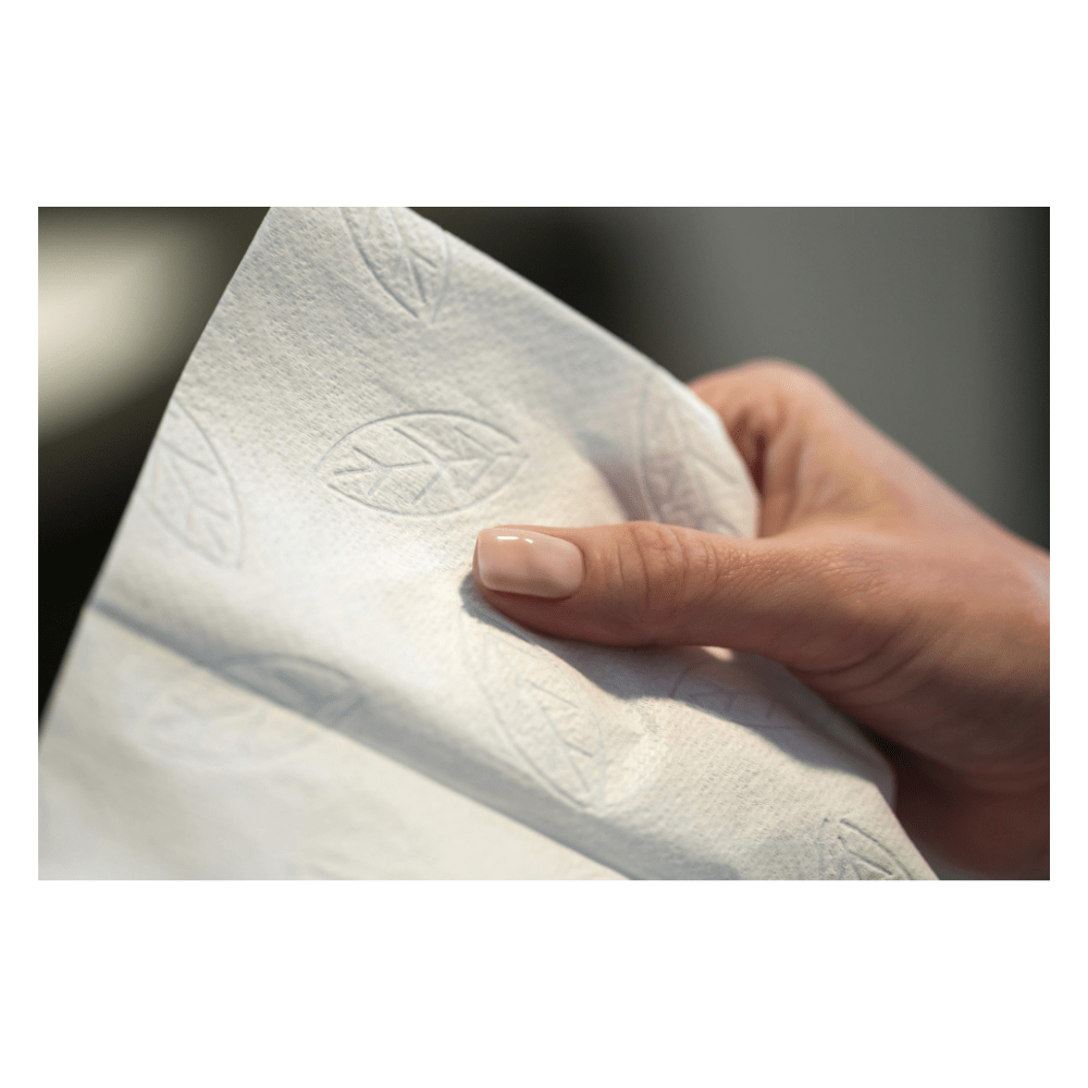 Une main tient une serviette en papier Tork 100278 de grande qualité, extra-douce, à motif en zigzag, gamme Premium H3, à deux couches, gaufrée d’un motif de feuilles. Le pouce et l’index enserrent cet accessoire d’hygiène de la marque TORK – Essity Professional Hygiene Germany GmbH. L’arrière-plan est flou, ce qui met en valeur l’élégance de la serviette.