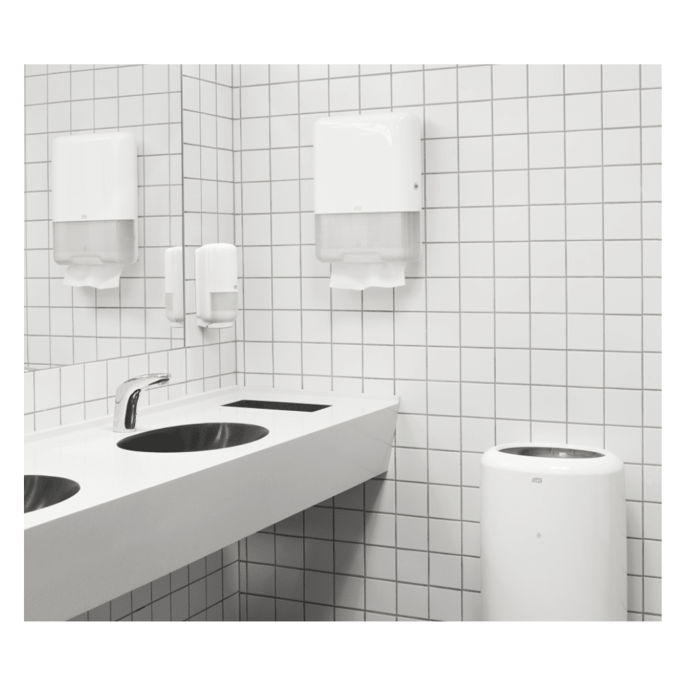 Des toilettes publiques modernes sont équipées d'un lavabo blanc doté de deux robinets, d'un mur carrelé de blanc et d'un distributeur TORK Tork 100278 de serviettes en papier extra-douces en zigzag Premium H3, 2 couches | Carton (15 paquets), contenant des serviettes en papier haut de gamme à deux couches. Complété par un distributeur de savon et une poubelle, l'espace est propre et épuré.