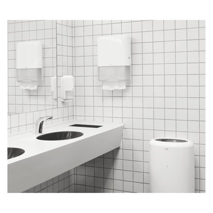 Des toilettes publiques modernes sont équipées d'un lavabo blanc doté de deux robinets, d'un mur carrelé de blanc et d'un distributeur TORK Tork 100278 de serviettes en papier extra-douces en zigzag Premium H3, 2 couches | Carton (15 paquets), contenant des serviettes en papier haut de gamme à deux couches. Complété par un distributeur de savon et une poubelle, l'espace est propre et épuré.