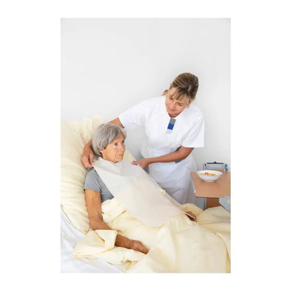 Une infirmière ajuste la couverture pour une femme âgée assise droite dans un lit d'hôpital. Les deux semblent attentives et concentrées. Un plateau avec un bol et Vala® Fit Tape Protection vestimentaire à usage unique est visible.