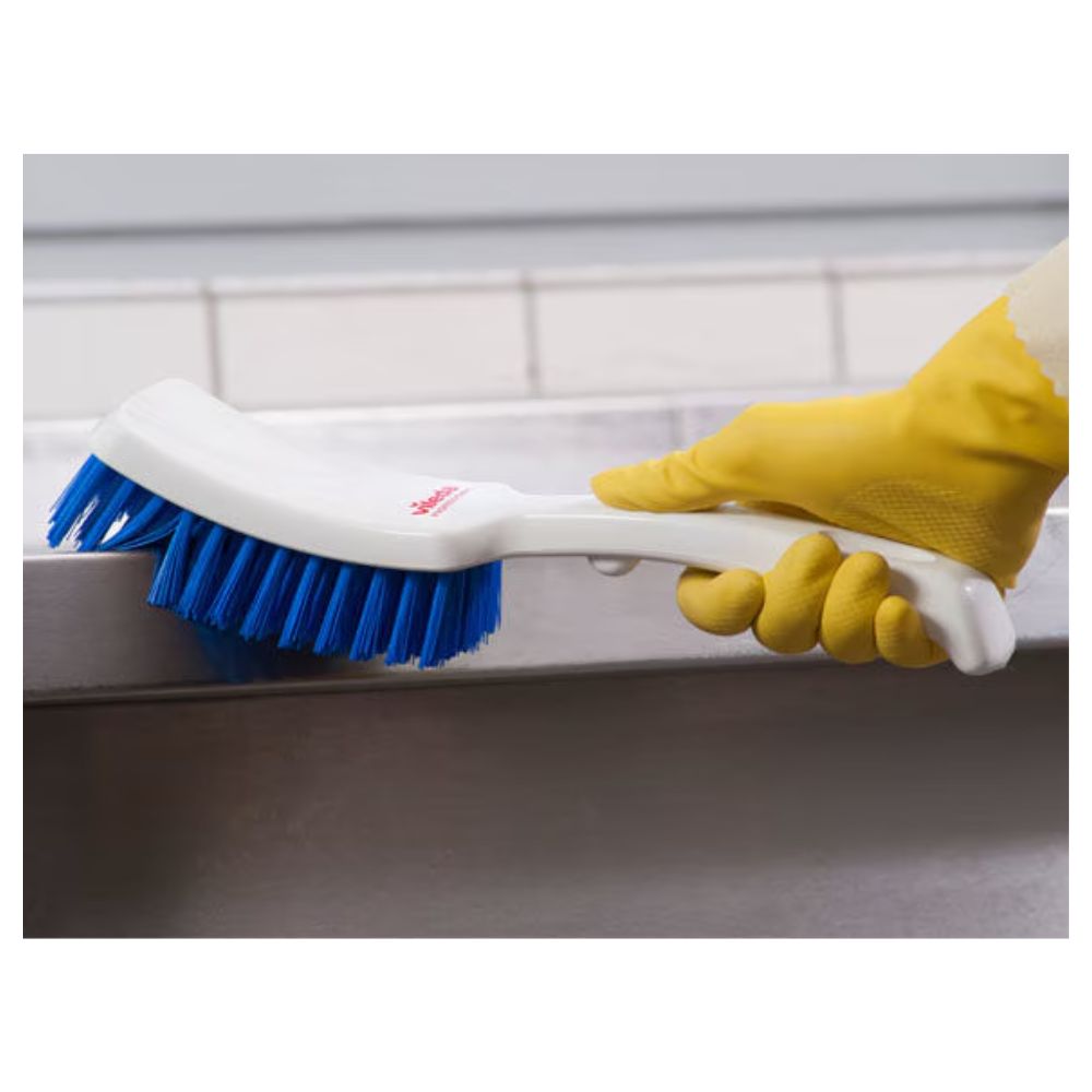 Une personne portant des gants en caoutchouc jaunes frotte une surface grise avec une brosse à manche court bleue Vileda Professional de 30 cm de Vileda GmbH, tandis qu'un mur carrelé offre un arrière-plan neutre.