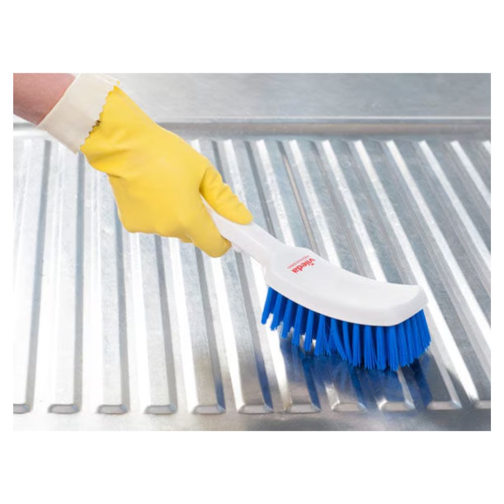 Une personne portant un gant en caoutchouc jaune utilise une brosse à manche court bleue Vileda Professional de 30 cm | Paquet (1 pièce) de Vileda GmbH avec des poils de haute qualité pour nettoyer une surface métallique rainurée.