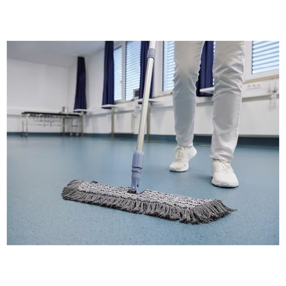 Une personne en chaussures et pantalons blancs nettoie avec un Vileda GmbH Professional Swep Duo r-MicroCombi Mop - 50 cm un sol bleu dans une pièce aux stores fermés et aux rideaux bleus. À proximité se trouvent une table pliante et un radiateur sous les fenêtres.