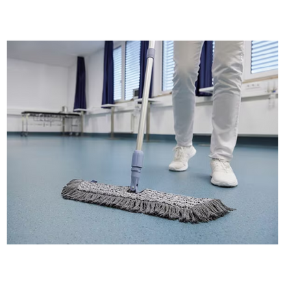 Une personne en chaussures et pantalons blancs nettoie avec un Vileda GmbH Professional Swep Duo r-MicroCombi Mop - 50 cm un sol bleu dans une pièce aux stores fermés et aux rideaux bleus. À proximité se trouvent une table pliante et un radiateur sous les fenêtres.
