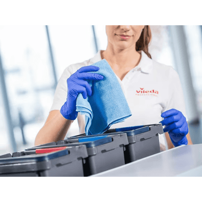 Une personne portant une chemise blanche et des gants bleus tient un chiffon en microfibre Vileda Professional r-MicroTuff Swift bleu de Vileda GmbH au-dessus d'une rangée de conteneurs avec des couvercles de différentes couleurs; la scène illustre la tâche de nettoyage efficace à l'aide du système à quatre couleurs pour l'organisation.