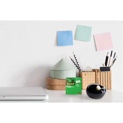 Un bureau avec un ordinateur portable fermé, 3M Scotch™ Magic Tape, un distributeur de ruban de table noir Scotch™ Karim Rashid de 3M Deutschland GmbH, des gobelets avec des stylos et crayons, des boîtes rondes empilées et trois notes autocollantes pastel sur un mur blanc.