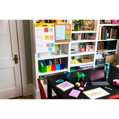 Dans un bureau à domicile se trouve une table noire avec des fournitures de bureau, une tablette et des carnets. À l'arrière-plan, il y a des étagères avec des livres et des classeurs ainsi qu'une liste de tâches sur un tableau blanc avec des Post-it® Super Sticky Notes (76x127 mm, 90 feuilles/bloc, 100% PEFC) de 3M.