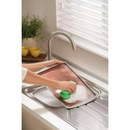 Quelqu'un frotte une grande plaque de cuisson sale avec l'éponge de nettoyage Scotch-Brite® Classic de 3M Deutschland GmbH sous un robinet dans une cuisine lumineuse avec des stores blancs, une plante et des bols jaunes empilés en arrière-plan.