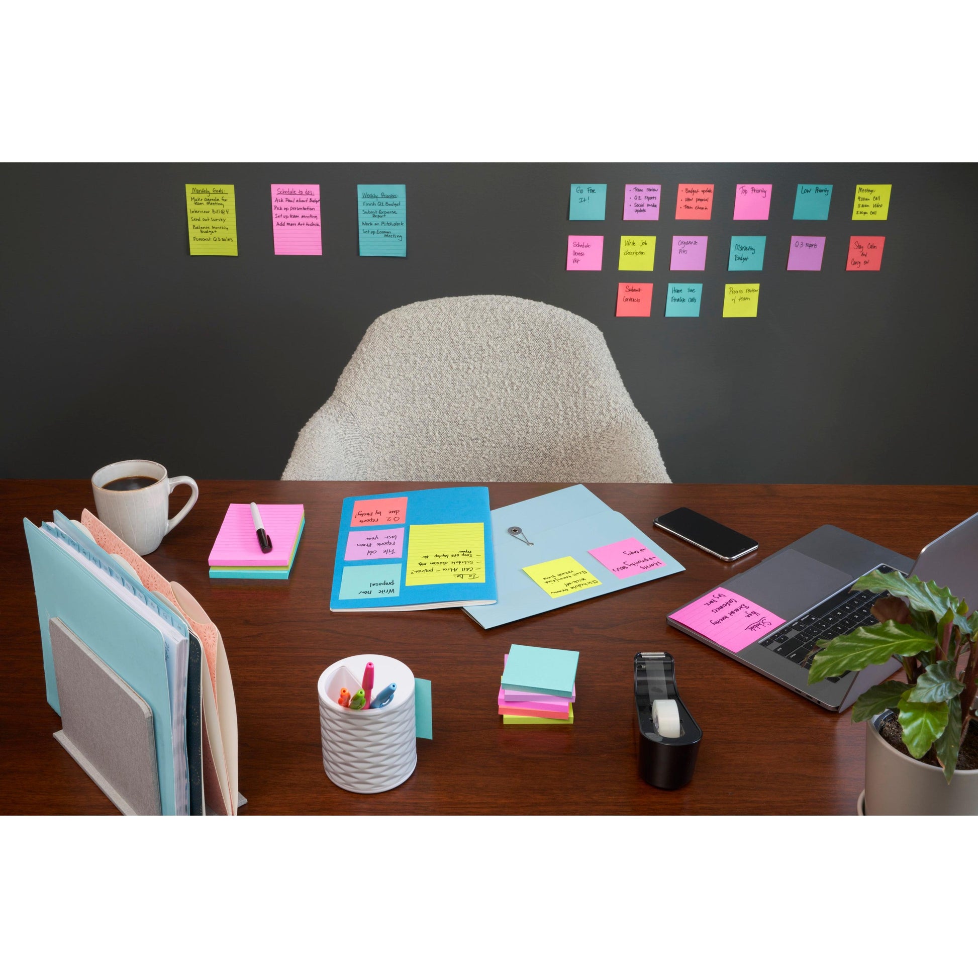Ein aufgeräumter Schreibtisch mit Laptop, Kaffee, Stiften und Büroutensilien steht vor einer Wand, die mit Reihen von 3M Deutschland GmbH Post-it® Super Sticky Notes (47,6mm x 47,6mm, 90 Blatt/Block, 100% PEFC) bedeckt ist; ein leerer Stuhl steht hinter dem Schreibtisch.