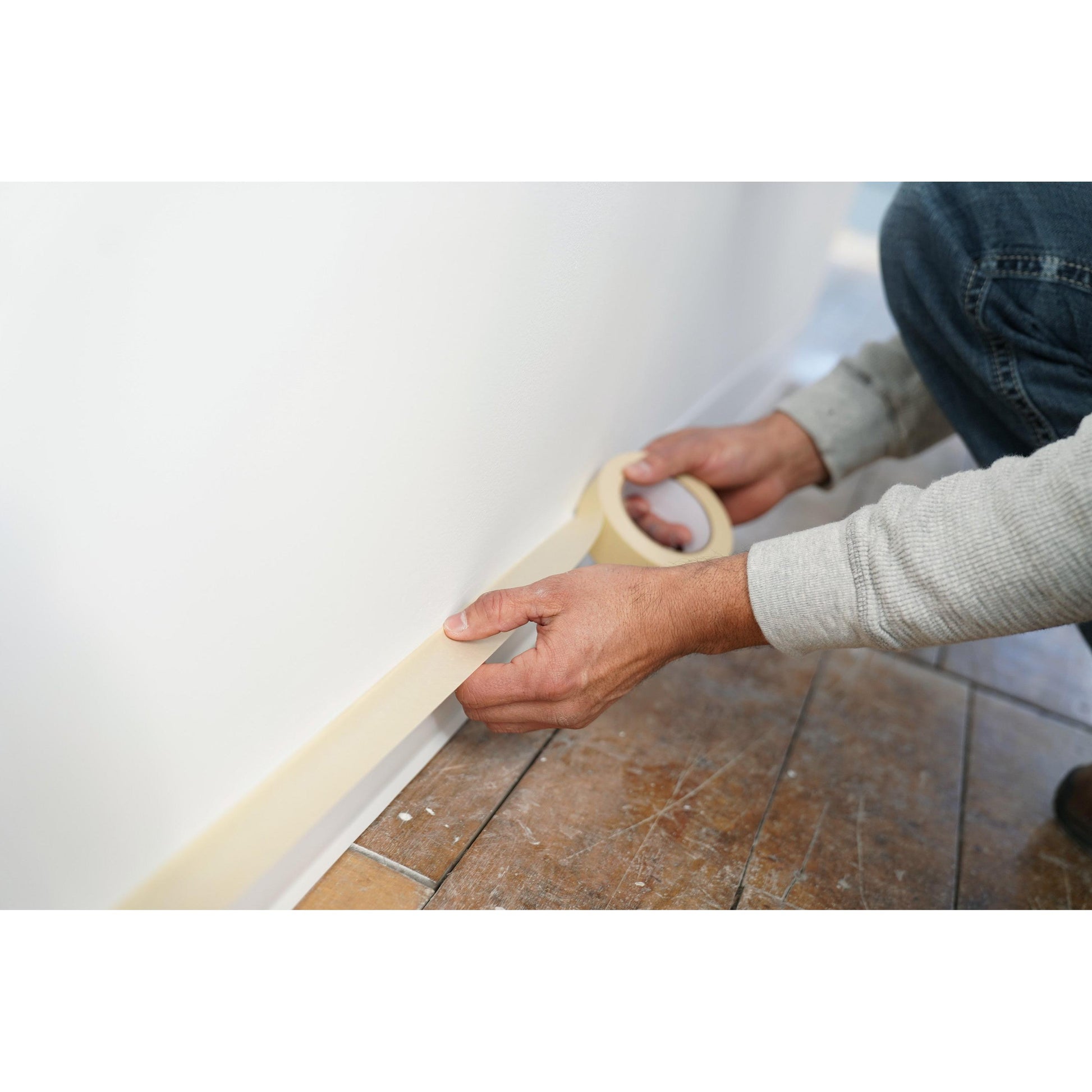 Jemand klebt Scotch® Abdeckband Classic (48 mm x 50 m, 3M Deutschland GmbH) entlang der Unterseite einer weißen Wand an der Sockelleiste und bereitet den Anstrich über einem leicht abgenutzten braunen Holzboden mit Farbspritzern vor.