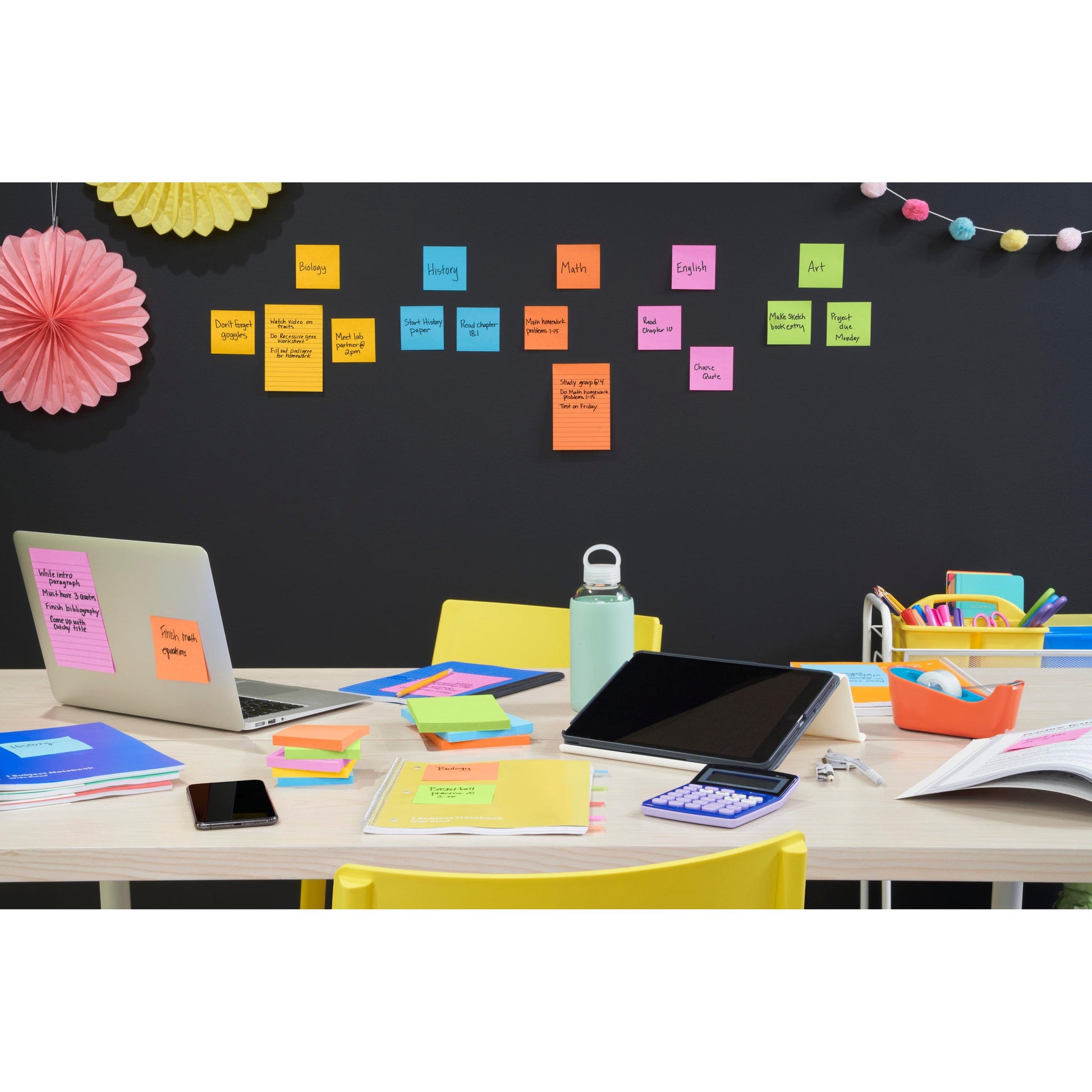 Ein Schreibtisch mit Laptop, Tablet, Taschenrechner, Telefon, Wasserflasche und bunten Schreibwaren. Dahinter ordnen Post-it® Super Sticky Notes (76x127mm) von 3M Deutschland GmbH die Schulfächer an der Wand, darüber hängen Dekorationen.