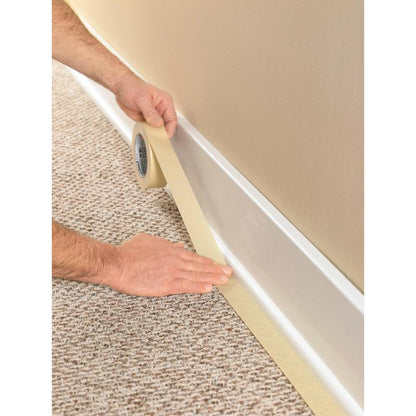 Une personne utilise le ruban de masquage Scotch® Classic, 24 mm x 50 m (70% PEFC) de 3M Deutschland GmbH, pour masquer la plinthe où la moquette rencontre le mur, afin de protéger le revêtement lors des travaux de peinture et de rénovation.