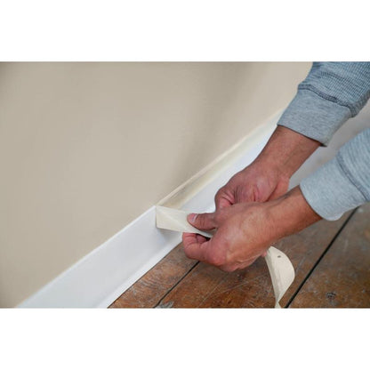 Lors de travaux de peinture et de rénovation, une bande de ruban de masquage Scotch® Classic (24 mm x 50 m, 1 rouleau/paquet, 70% PEFC) de 3M Deutschland GmbH est retirée du dessus d'une plinthe blanche le long d'un mur beige avec un sol en bois.