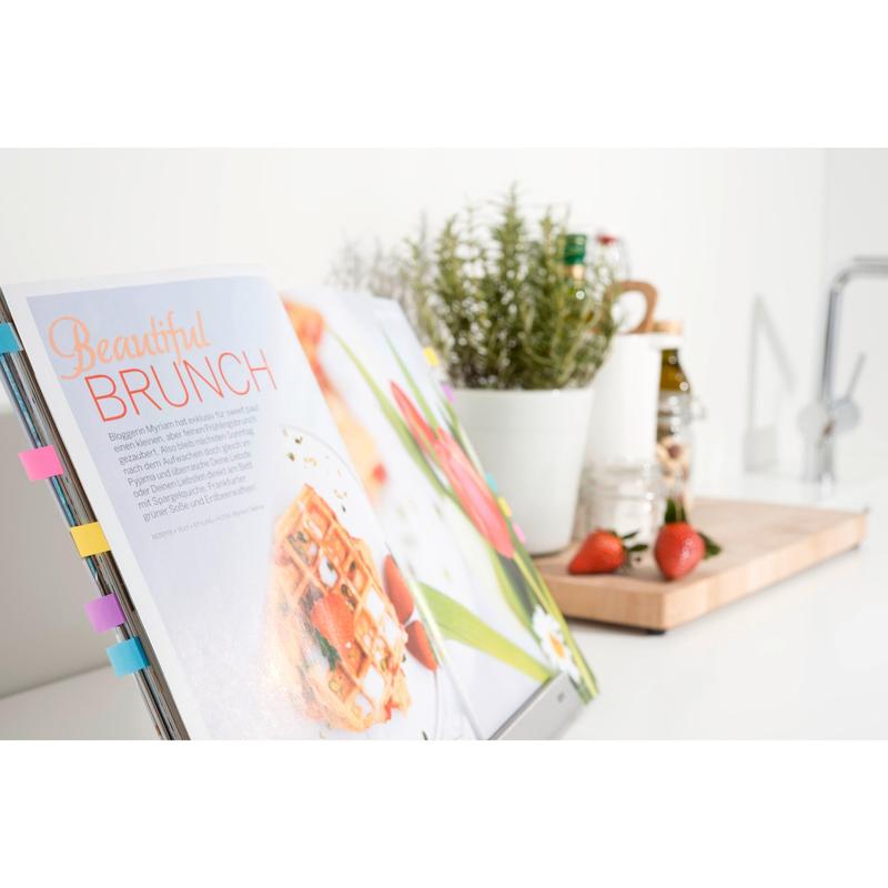 Un livre de cuisine étiqueté avec des Post-it® Index Signets Mini (11,9 x 43,2 mm, 100/distributeur) de 3M Deutschland GmbH montre une recette de brunch ; derrière se trouvent un pot d'herbes blanc, des fraises sur une planche et des ustensiles de cuisine sur un comptoir ensoleillé.