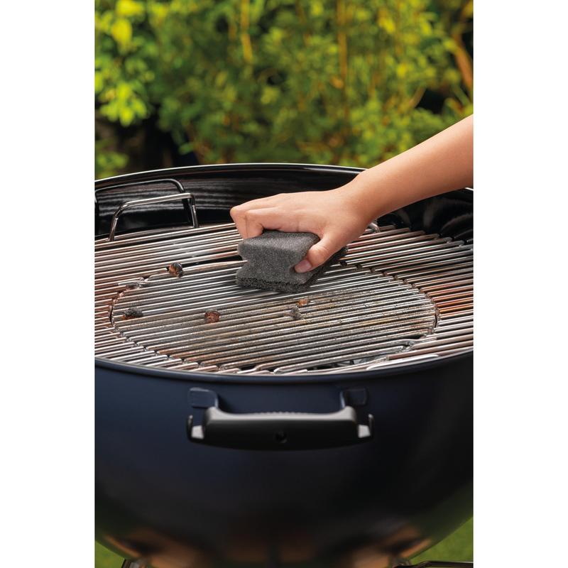 Une main nettoie la grille en métal d'un barbecue rond au charbon de bois avec un tampon à manche Scotch-Brite® (1 pièce par paquet) de 3M Deutschland GmbH. Le feuillage vert est flou en arrière-plan.