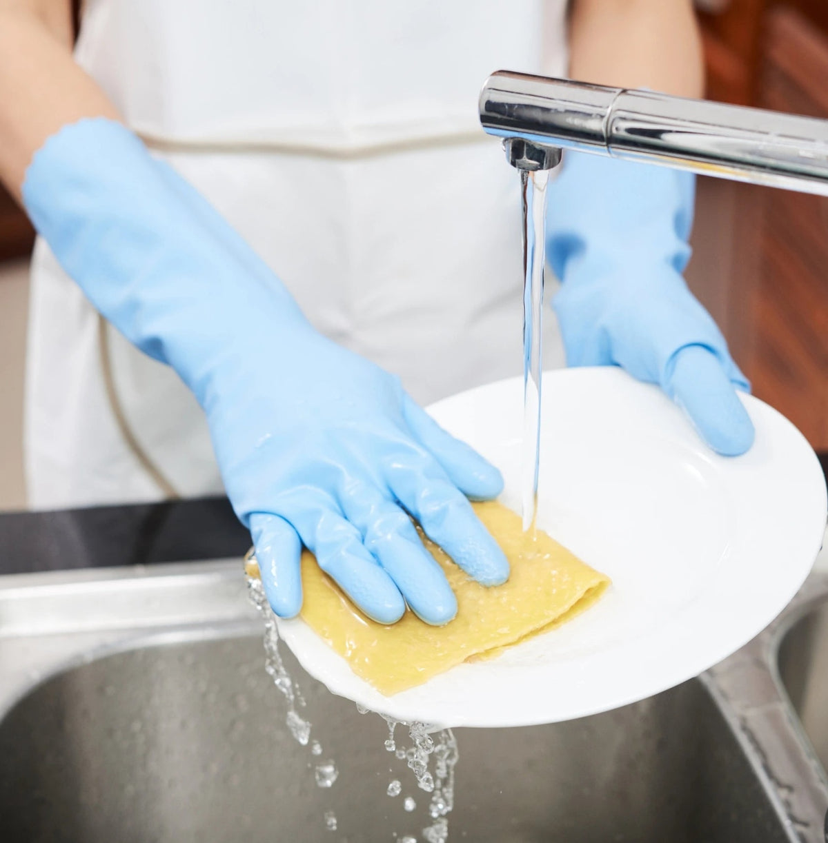 Hände in blauen Haushaltshandschuhen spülen einen weißen Teller mit Schwammtuch unter dem Wasserhahn